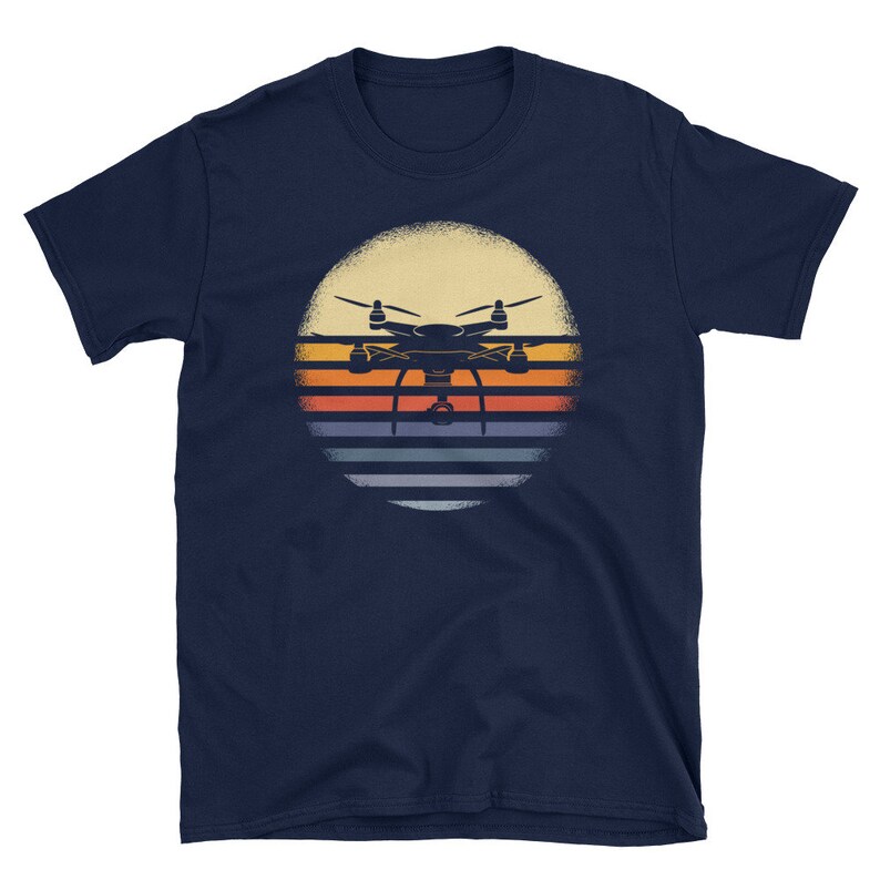 May include: A navy blue t-shirt with a vintage-style graphic of a drone flying over a sunset. The graphic features a silhouette of a drone with a camera, set against a background of orange, yellow, and blue stripes.