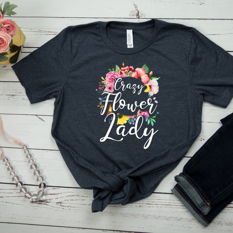 May include: Dark navy blue t-shirt with the text "Crazy Flower Lady" in white script, surrounded by a colorful floral wreath. The shirt is made of a soft material and is paired with dark blue jeans.