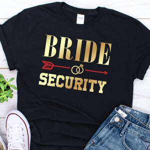 May include: Black t-shirt with gold text that reads "Bride Security" and a red arrow with gold rings.