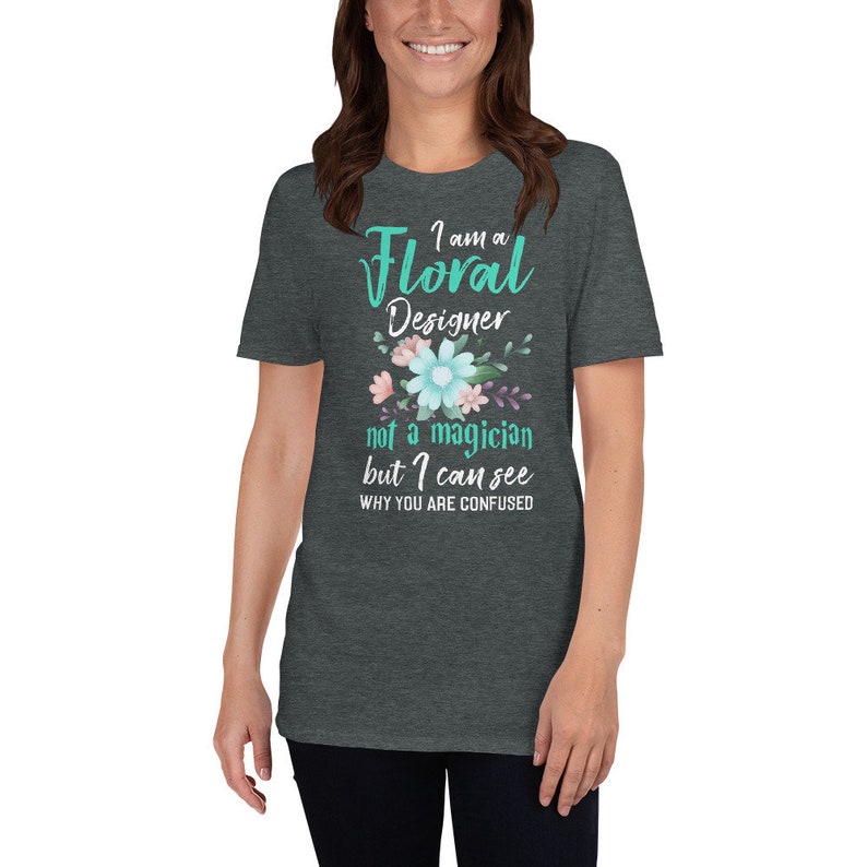 May include: A dark heather grey t-shirt with a teal green graphic that says "I am a Floral Designer not a magician but I can see why you are confused". The graphic is surrounded by a floral design with pink, blue, and green flowers.