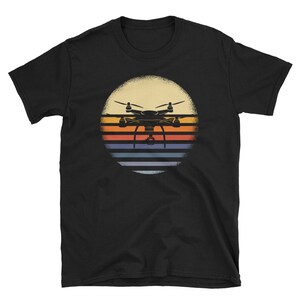 May include: Black t-shirt with a retro-style graphic of a drone flying over a sunset. The graphic features a silhouette of a drone with a camera, set against a background of orange, yellow, and blue stripes.