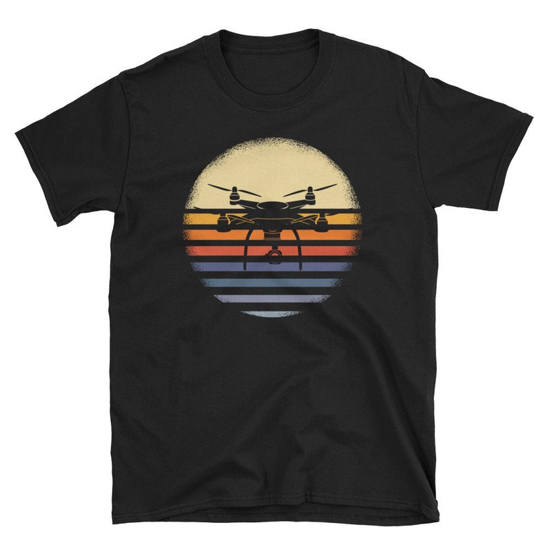 May include: Black t-shirt with a retro-style graphic of a drone flying over a sunset. The graphic features a silhouette of a drone with a camera, set against a background of orange, yellow, and blue stripes.