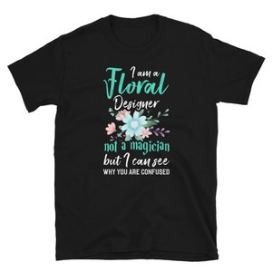 May include: Black t-shirt with a teal green graphic that says "I am a Floral Designer not a magician but I can see why you are confused". The graphic also includes a floral design with pink, blue, and purple flowers.