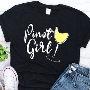 以下が含まれることがあります： 「Pinot Girl!」という白い文字と黄色のワイングラスのグラフィックが描かれた黒いTシャツです。