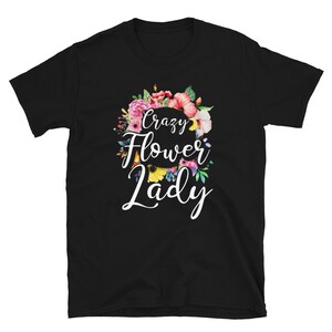May include: Black t-shirt with a floral wreath design and the words "Crazy Flower Lady" in white script. The wreath features pink, red, and yellow flowers with green leaves. A casual tee for flower enthusiasts.