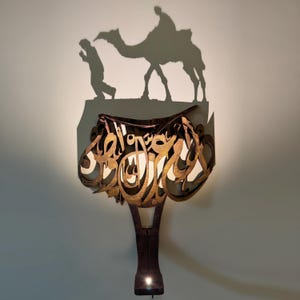 Puede incluir: Una lámpara de pared decorativa con un diseño único. La lámpara presenta un marco de metal marrón con caligrafía árabe y una luz cálida. La sombra proyectada por la lámpara representa un camello y figuras, añadiendo un toque cultural.
