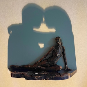 Op de afbeelding: Een houten sculptuur van een liggende figuur met de schaduw van een koppel dat elkaar omarmt erachter. Het beeld is donkerbruin en heeft een ruw, getextureerd oppervlak.