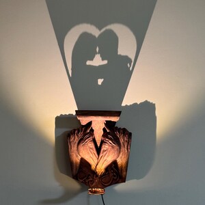Op de afbeelding: Een wandlamp met een houten voet en twee gebeeldhouwde handen. De lamp werpt een schaduw van een koppel dat elkaar omhelst in de vorm van een hart op de muur.