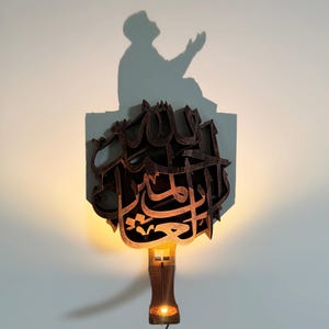 Può includere: Un'opera d'arte da parete in legno con calligrafia araba, illuminata dal basso. Il design proietta l'ombra di una persona in preghiera sul muro. L'opera è marrone ed emana una luce calda.