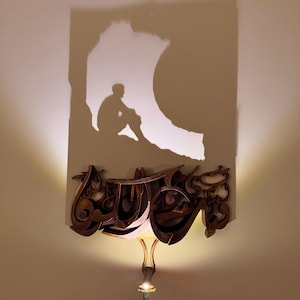 May include: A shadow of a person sitting on a rock is projected onto a wall. The shadow is cast by a light fixture that is shaped like Arabic calligraphy. The light fixture is made of brown wood and has a light bulb that is turned on.
