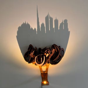 May include: A wooden wall sconce with a light fixture and a shadow of a city skyline behind it. The sconce is made of carved wood and has a warm, inviting glow.