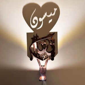 May include: A wooden wall-mounted light fixture with a heart-shaped cutout that casts a shadow of the Arabic word "سَوْرَ" on the wall. The light fixture is designed to cast a shadow of the Arabic word "عَلَيْكُمُ السَّلَامُ" when lit.