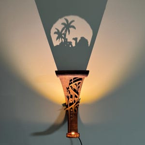 May include: A wooden wall sconce with intricate Arabic calligraphy designs. The sconce casts a shadow of palm trees and a camel on the wall.