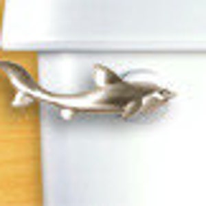 May include: A silver-colored shark-shaped lapel pin. The pin is detailed with fins, a tail, and an open mouth, and is attached to a white surface. The pin is approximately 1 inch long.
