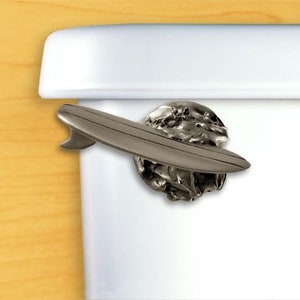 May include: A silver toilet paper holder shaped like a surfboard and a wave. The holder is mounted on a white toilet tank.