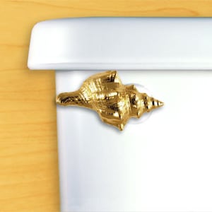 May include: Gold seashell toilet paper holder. The seashell is a realistic design and is mounted on a white toilet tank.