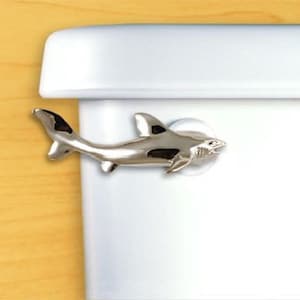 May include: A silver-colored shark-shaped toilet seat lifter. The shark has black accents on its fins and tail and is attached to the toilet seat with a clear suction cup. The lifter is designed to be decorative and functional.