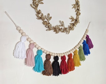 May include: A rainbow tassel garland with wooden beads hangs on a white wall. The garland features tassels in various colors, including white, pink, turquoise, brown, black, red, yellow, and green.