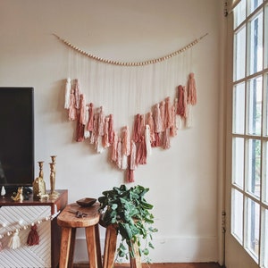 May include: A wall hanging with a wooden bead garland and cascading tassels in shades of pink, peach, and white. The tassels are suspended from strings, creating a decorative, bohemian-style accent. The wall hanging is displayed above a wooden stool and a cabinet.