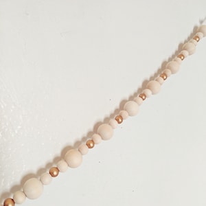 Peut inclure: Une guirlande de perles en bois avec des accents dor&eacute;s. Les perles sont enfil&eacute;es sur une ficelle blanche. La guirlande est suspendue &agrave; un mur blanc.