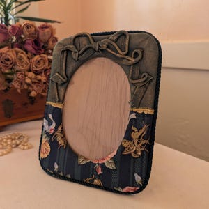 May include: An oval picture frame featuring a dark blue fabric base with a floral and cherub print, accented by a gold-toned fabric top section with a decorative bow. The frame is edged with dark blue twisted rope trim and has a light wood center.