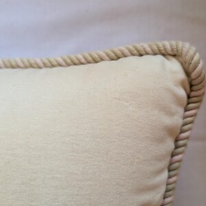 May include: A square throw pillow in a solid light beige color featuring a thick, twisted rope trim along the edge. The fabric has a smooth, matte texture with a subtle, small indentation visible on the surface of the pillow cover material.