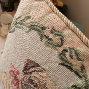 May include: A square needlepoint pillow featuring a tan pug with a pink bow around its neck. The pillow has a cream-colored background with green floral vine patterns and is finished with a thick, twisted beige rope trim along the edge.
