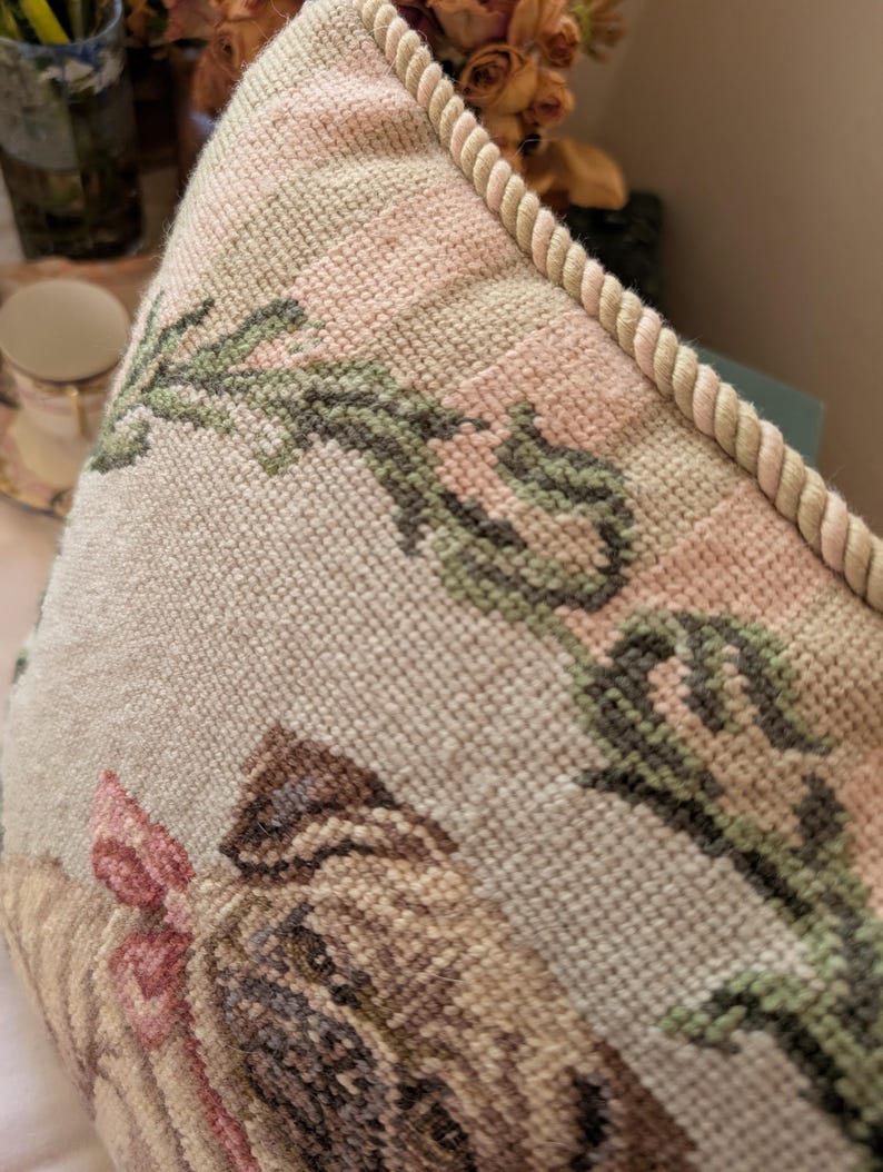 May include: A square needlepoint pillow featuring a tan pug with a pink bow around its neck. The pillow has a cream-colored background with green floral vine patterns and is finished with a thick, twisted beige rope trim along the edge.