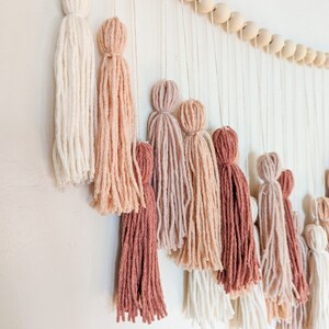 May include: A wall hanging featuring a series of yarn tassels in varying shades of white, pink, and brown. The tassels are suspended from a string of wooden beads, creating a decorative and textured design.