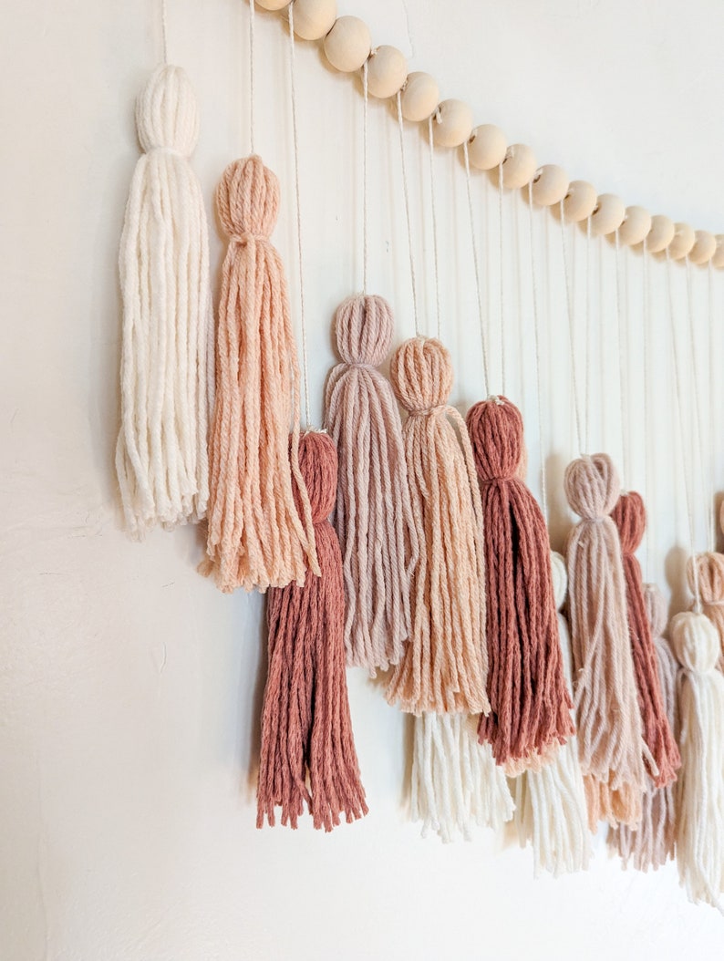 May include: A wall hanging featuring a series of yarn tassels in varying shades of white, pink, and brown. The tassels are suspended from a string of wooden beads, creating a decorative and textured design.