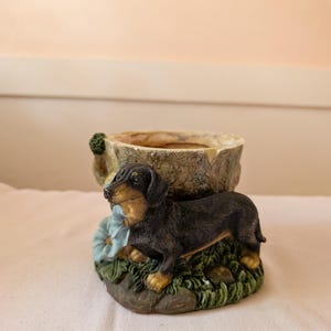 May include: A decorative planter featuring a dachshund dog figurine. The dog is black and tan, positioned on a base with green grass, blue flowers, and a stone-like planter. The planter has a rough, textured appearance.