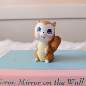 May include: A small ceramic squirrel figurine with big blue eyes and a brown and white body. The figurine is standing on a book with the title "Mirror, Mirror on the Wall".