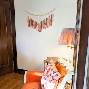 May include: A wall hanging with a series of tassels in shades of beige, peach, and brown, suspended from a string of wooden beads. The tassels are arranged in a descending V-shape. The wall hanging is displayed on a white wall.