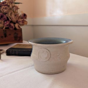 May include: A small, off-white ceramic pot with a blue interior rim and a circular embossed design. The pot has a slightly textured surface and a rounded shape. The pot is sitting on a white tablecloth.