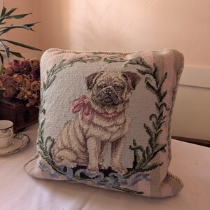 May include: A square needlepoint throw pillow featuring a tan pug dog wearing a pink bow around its neck. The pug is centered within a green leafy wreath design on a light-colored background with a thick, twisted rope-style border trim.
