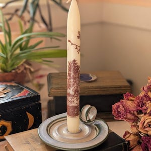 May include: A tall, cream-colored candle with a brown landscape design stands in a ceramic candle holder. The holder is a light green and gray with a spiral design. The candle sits on a stack of books.