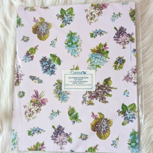K&ouml;nnte beinhalten: Eine Packung Geschenkpapier mit viktorianischen Blumenstr&auml;u&szlig;en, die ein Blumenmuster mit blauen, violetten und gr&uuml;nen Blumen auf einem hellrosa Hintergrund zeigt. Die Packung enth&auml;lt zwei 61 cm x 76 cm gro&szlig;e Geschenkpapierb&ouml;gen. Der Text auf der Packung lautet "Current Victorian Nosegays Gift Wrap Code 18161-7".