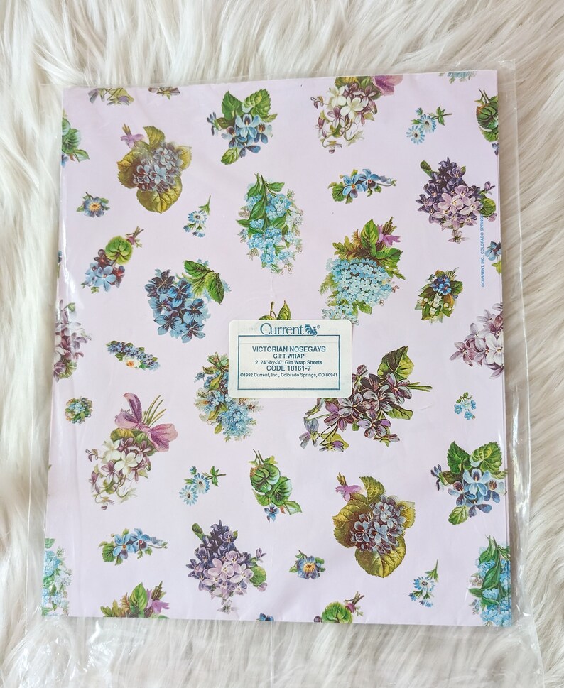 K&ouml;nnte beinhalten: Eine Packung Geschenkpapier mit viktorianischen Blumenstr&auml;u&szlig;en, die ein Blumenmuster mit blauen, violetten und gr&uuml;nen Blumen auf einem hellrosa Hintergrund zeigt. Die Packung enth&auml;lt zwei 61 cm x 76 cm gro&szlig;e Geschenkpapierb&ouml;gen. Der Text auf der Packung lautet "Current Victorian Nosegays Gift Wrap Code 18161-7".