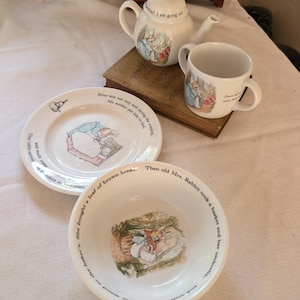 May include: A white ceramic tea set and bowl featuring Peter Rabbit illustrations and printed text. The set includes a teapot, a two-handled mug, a plate, and a bowl, all decorated with scenes of rabbits and excerpts from the classic storybook.
