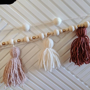 Peut inclure: Une guirlande avec des perles en bois et des accents dor&eacute;s, enfil&eacute;es avec des pompons blancs et des glands en laine dans des tons de rose, blanc et marron.
