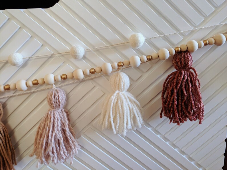 Peut inclure: Une guirlande avec des perles en bois et des accents dor&eacute;s, enfil&eacute;es avec des pompons blancs et des glands en laine dans des tons de rose, blanc et marron.