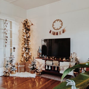 Peut inclure: Un salon d&eacute;cor&eacute; pour No&euml;l avec un grand sapin de No&euml;l fin et haut orn&eacute; de boules blanches et de lumi&egrave;res. Le sapin est surmont&eacute; d'une trompette dor&eacute;e. Un t&eacute;l&eacute;viseur est mont&eacute; au mur au-dessus d'un meuble de divertissement en bois avec une guirlande de glands et une couronne. Il y a deux petits sapins de No&euml;l sur le sol et une grande plante en pot au premier plan.