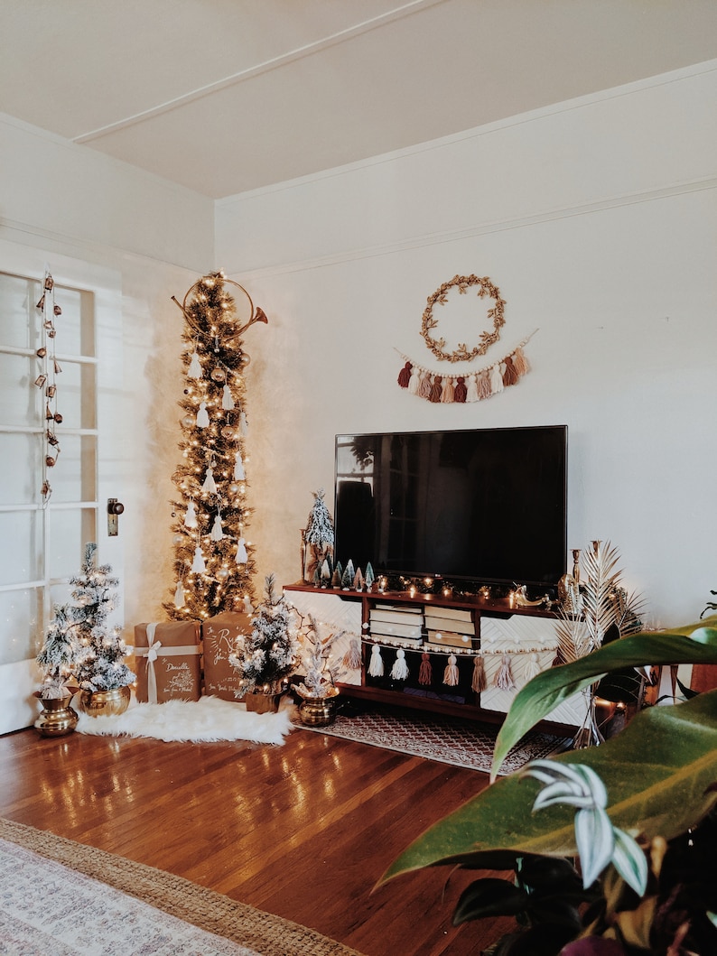 Peut inclure: Un salon d&eacute;cor&eacute; pour No&euml;l avec un grand sapin de No&euml;l fin et haut orn&eacute; de boules blanches et de lumi&egrave;res. Le sapin est surmont&eacute; d'une trompette dor&eacute;e. Un t&eacute;l&eacute;viseur est mont&eacute; au mur au-dessus d'un meuble de divertissement en bois avec une guirlande de glands et une couronne. Il y a deux petits sapins de No&euml;l sur le sol et une grande plante en pot au premier plan.