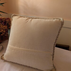 May include: A square off-white throw pillow featuring a smooth, solid-colored fabric front and back. The pillow is finished with a thick, twisted rope-style trim along all four edges, providing a textured border that frames the plain central panel.