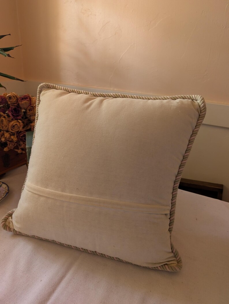 May include: A square off-white throw pillow featuring a smooth, solid-colored fabric front and back. The pillow is finished with a thick, twisted rope-style trim along all four edges, providing a textured border that frames the plain central panel.