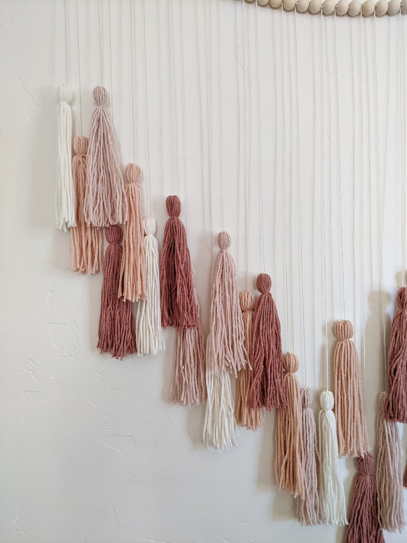 May include: A wall hanging featuring multiple yarn tassels in varying shades of pink, cream, and burgundy. The tassels are suspended from thin, vertical strings, creating a layered, textured effect. Wooden beads are visible at the top.