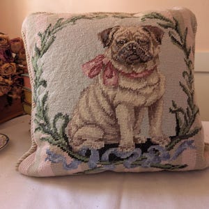 May include: A square throw pillow featuring a needlepoint design of a tan pug dog wearing a pink bow around its neck. The pug is centered within a green leafy wreath, with a blue ribbon draped across the bottom edge of the pillow cover.