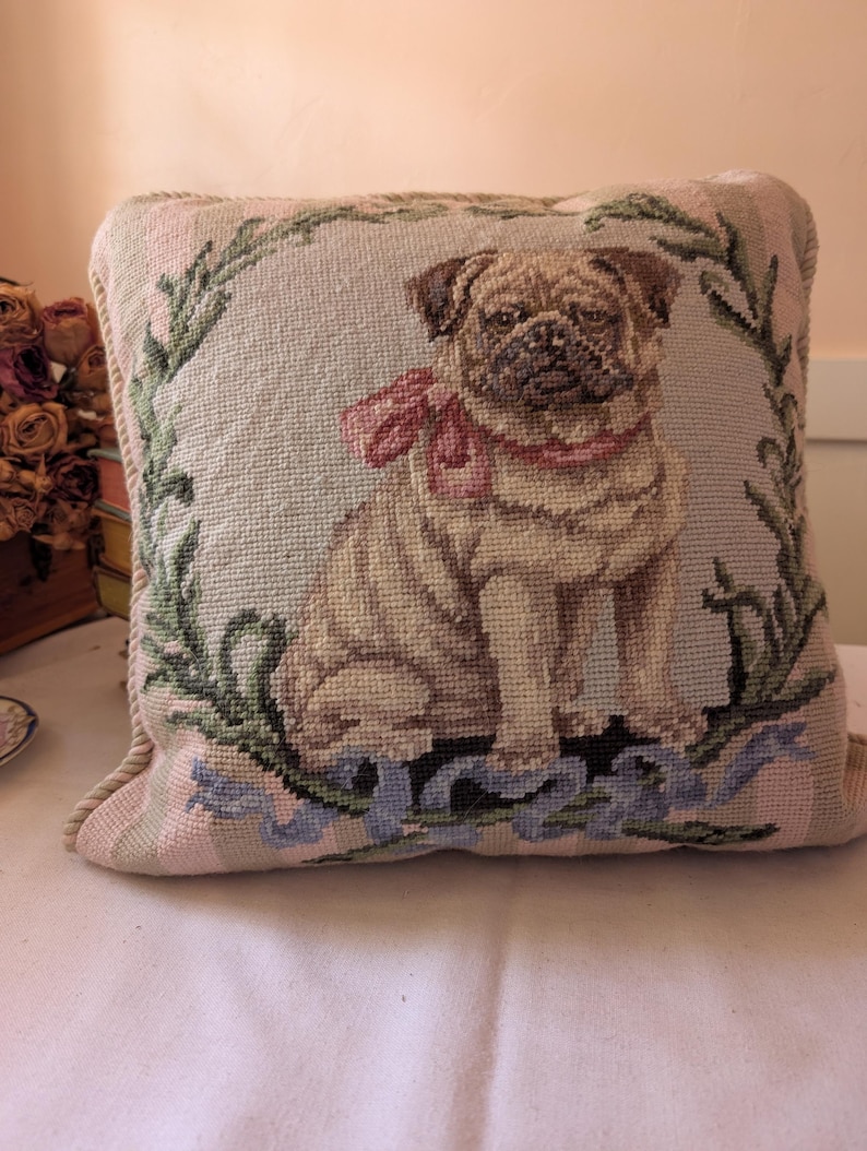 May include: A square throw pillow featuring a needlepoint design of a tan pug dog wearing a pink bow around its neck. The pug is centered within a green leafy wreath, with a blue ribbon draped across the bottom edge of the pillow cover.