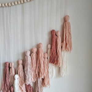 May include: A wall hanging featuring a cascade of yarn tassels in varying shades of pink, from deep rose to pale blush, suspended from a string of wooden beads. The tassels are arranged in a descending diagonal pattern against a white wall.