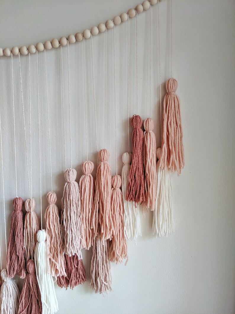 May include: A wall hanging featuring a cascade of yarn tassels in varying shades of pink, from deep rose to pale blush, suspended from a string of wooden beads. The tassels are arranged in a descending diagonal pattern against a white wall.